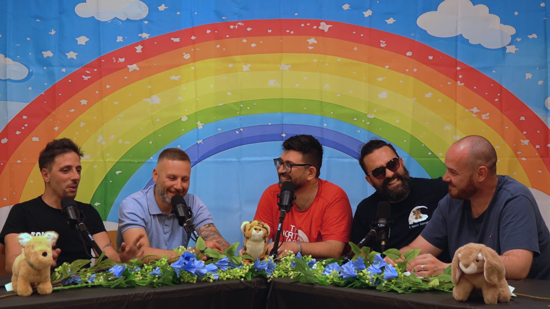 I conduttori del Podcast dei Papà al tavolo con microfoni, peluche e lo sfondo con arcobaleno